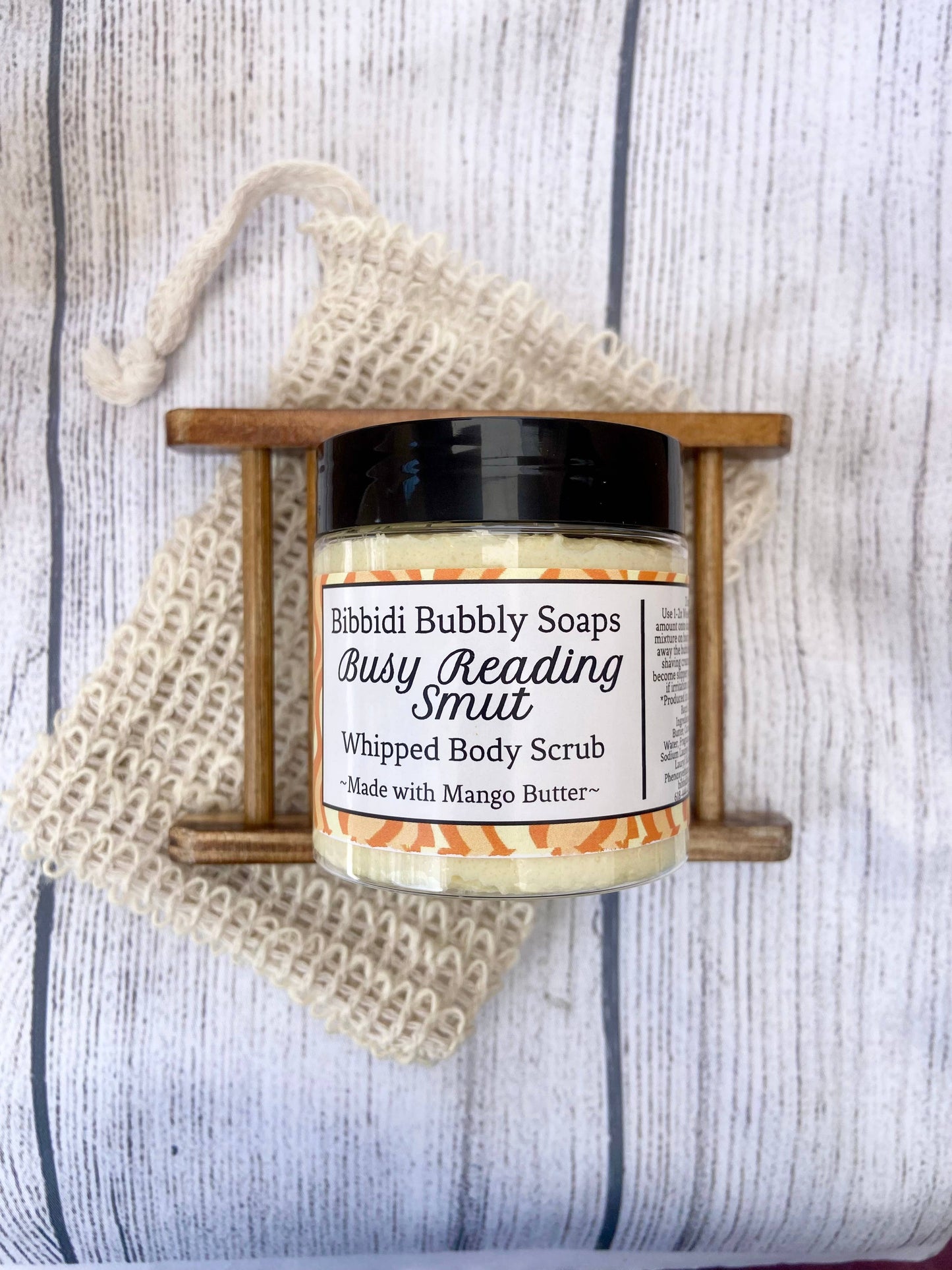Jar of 'Busy Reading Smut' whipped body scrub with a wooden holder on a textured surface