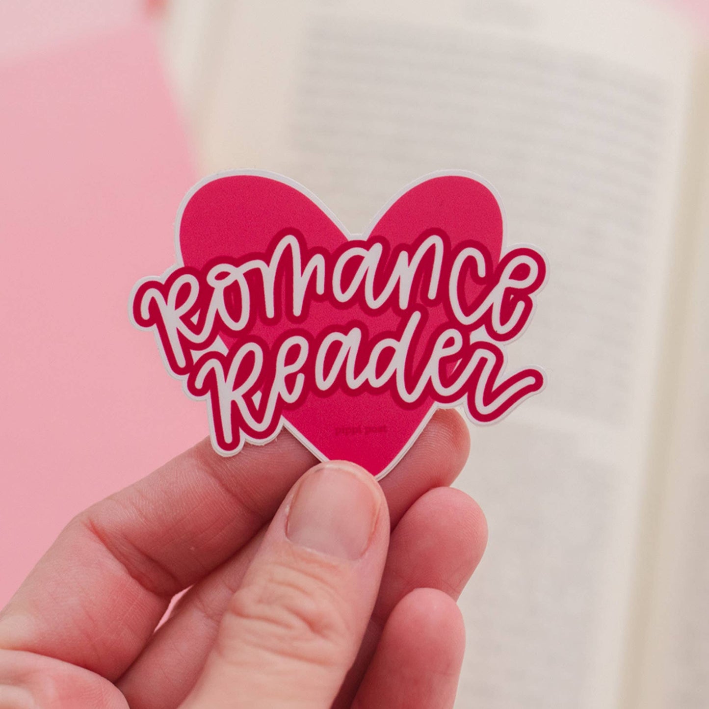 Heart-shaped sticker with 'Romance Reader' text held by a hand against a pink and beige background