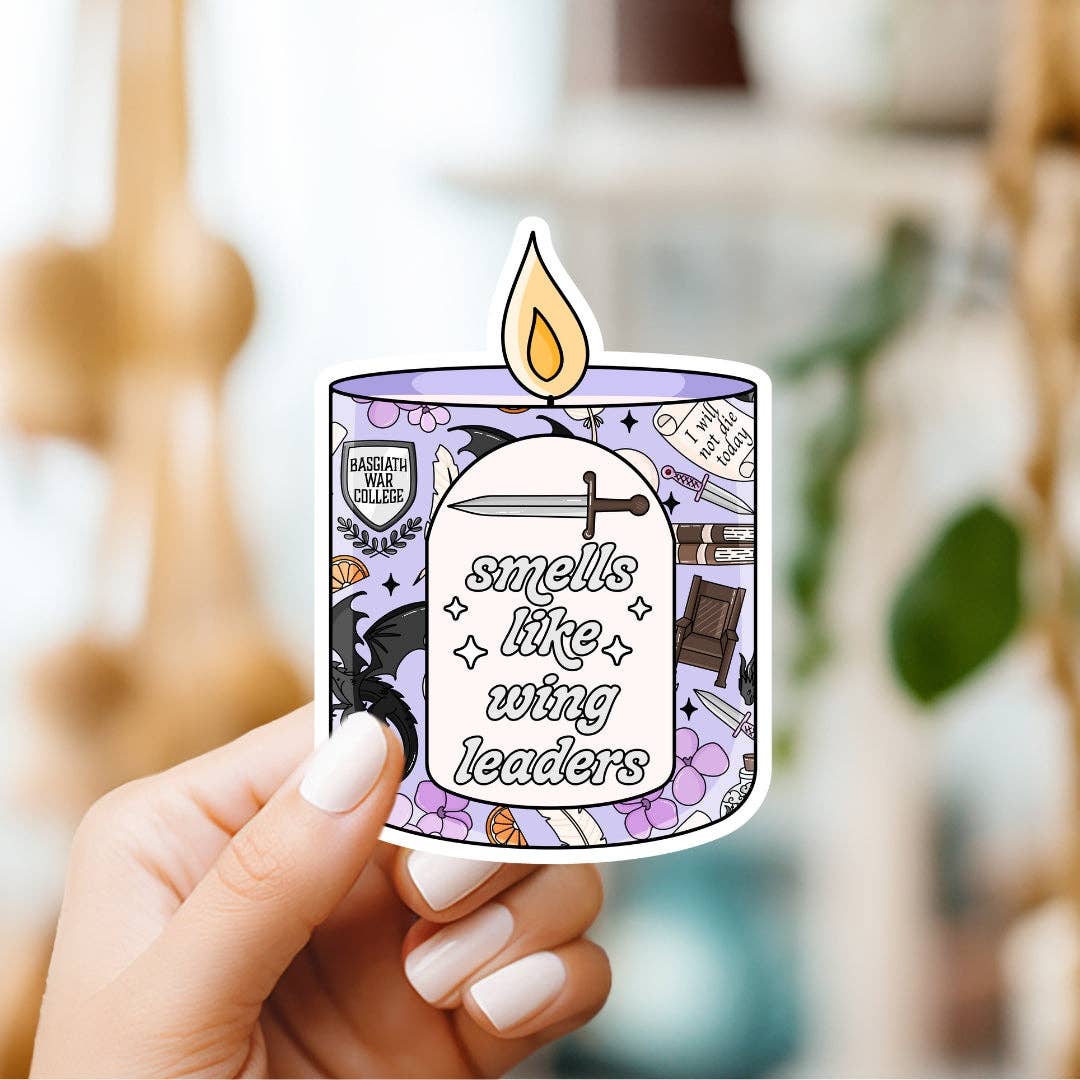 Hand holding a sticker with a candle design and text 'smells like wing leaders' against a blurred indoor background.