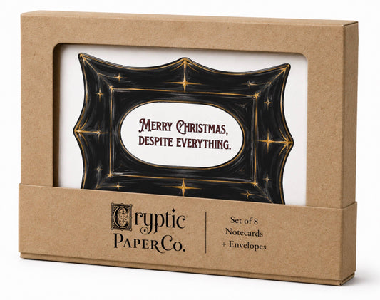 Cryptic Paper Co. Christmas greeting card set in a kraft paper box with a clear window, featuring a black card with gold stars and the text 'Merry Christmas, Despite Everything.