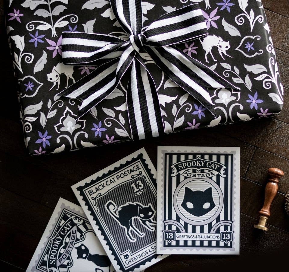 Halloween-themed wrapping paper and postcards featuring a black cat motif. A gift wrapped in black wrapping paper with white and purple floral and cat patterns is tied with a black and white striped ribbon.