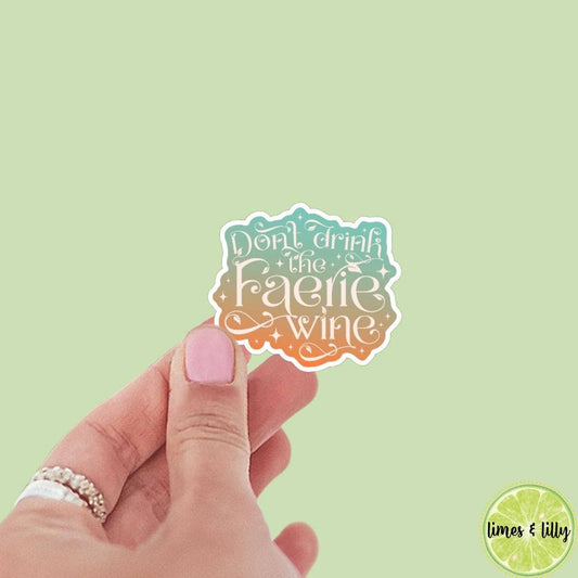 Hand holding a sticker with 'Don't drink the faerie wine' text against a light green background