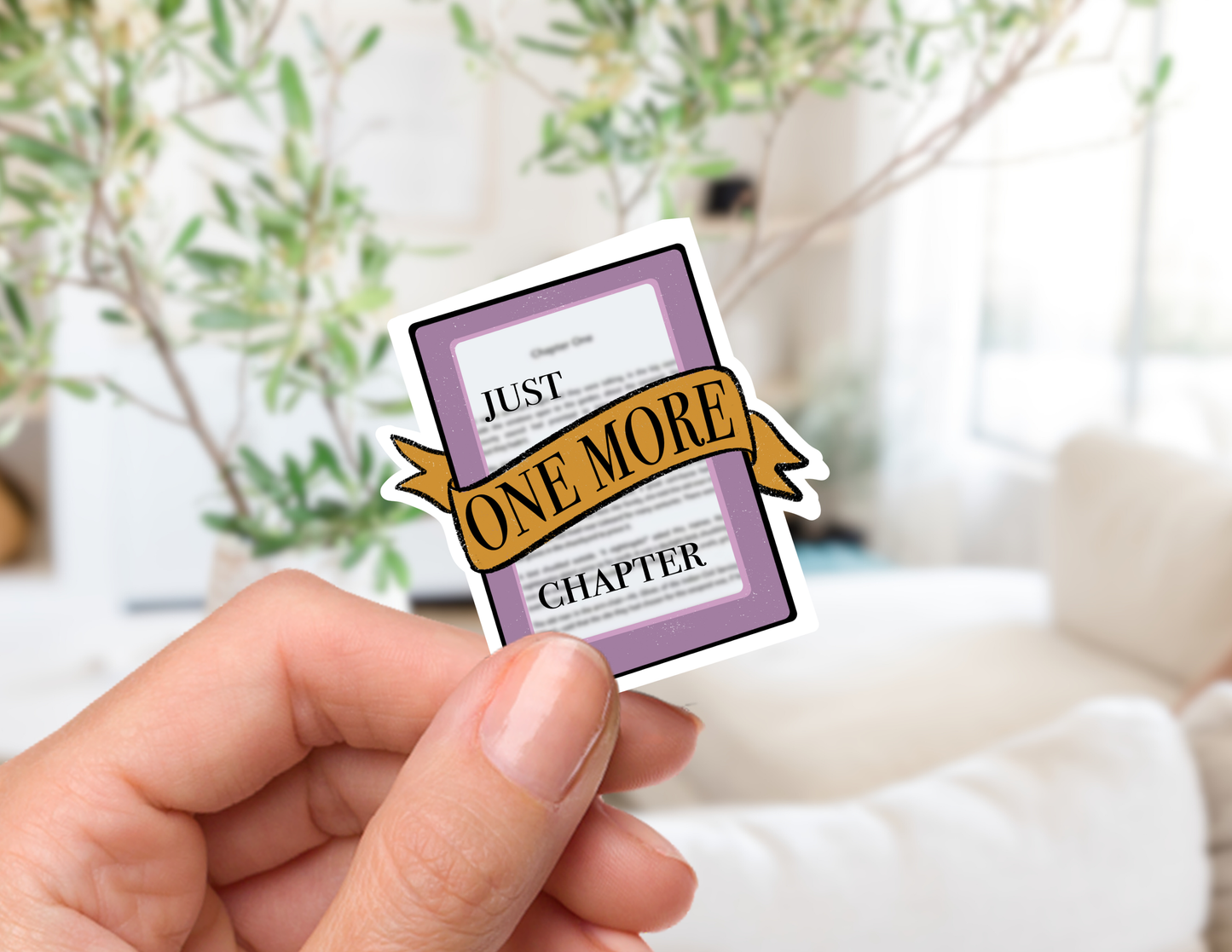A hand holds a die-cut sticker shaped like a book chapter, featuring a purple border, white page background, and a gold ribbon banner that reads 'JUST ONE MORE CHAPTER'. The background is a softly blurred indoor setting with a sofa and greenery.