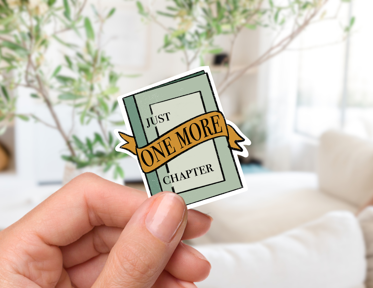 A hand holds a die-cut sticker shaped like a book cover with the text 'JUST ONE MORE CHAPTER' on a ribbon, set against a softly blurred indoor background with plants and a sofa.