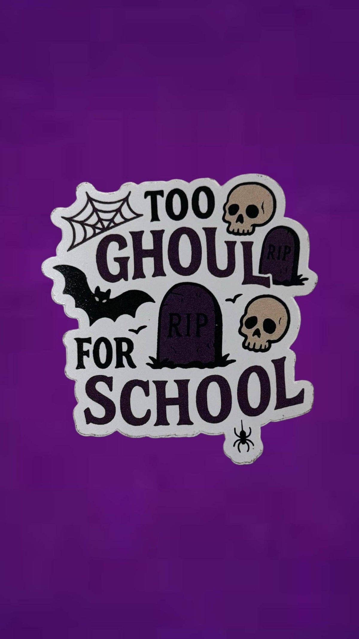 A Halloween-themed die-cut sticker with the text 'TOO GHOUL FOR SCHOOL' in dark purple lettering with a white outline. The design includes spooky illustrations of tombstones, skulls, a bat, a spider, and a spiderweb on a solid purple background.