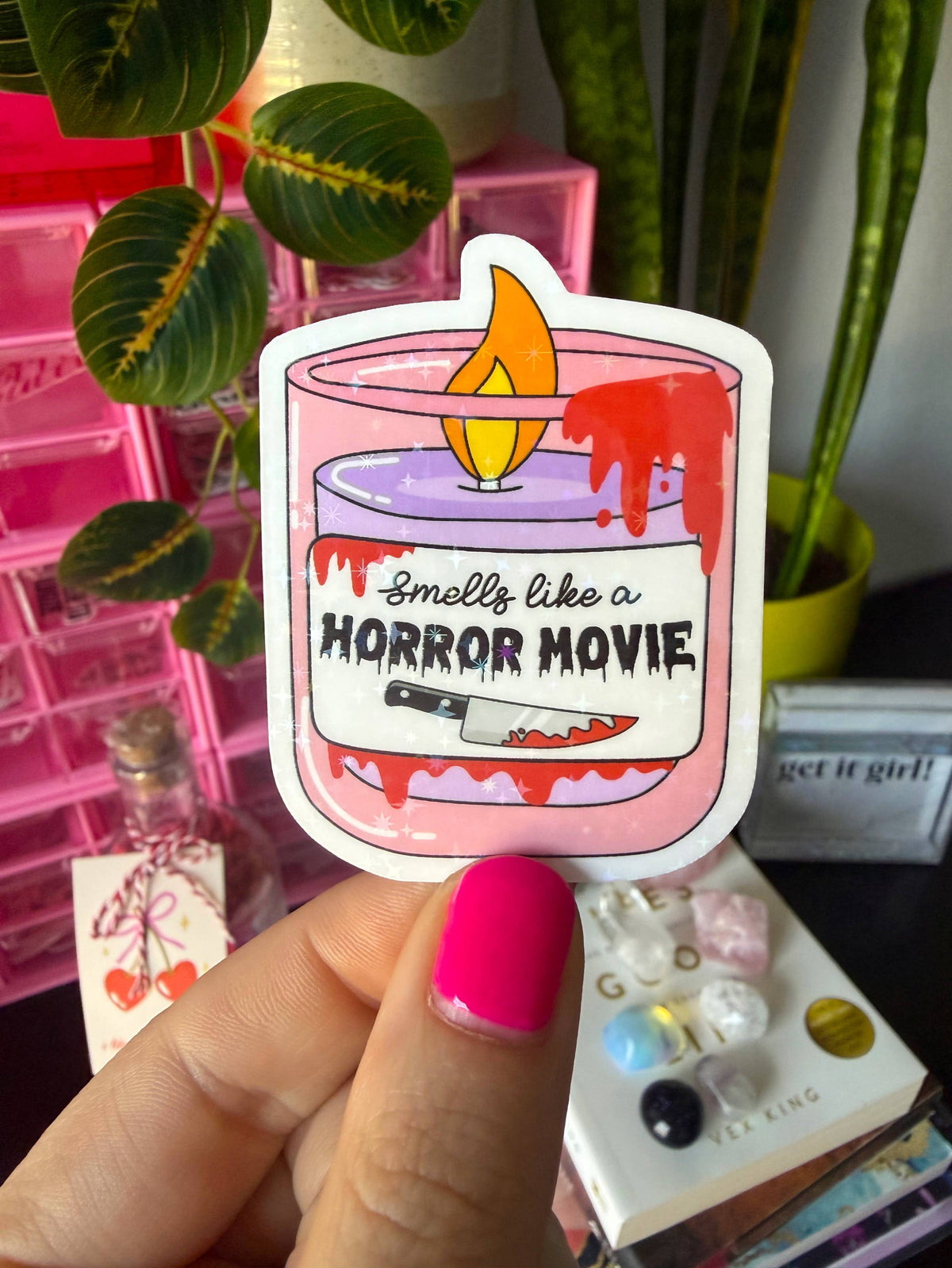 A hand with bright pink nail polish holds a die-cut sticker featuring a pink candle with dripping red wax, a yellow flame, and the text 'Smells like a HORROR MOVIE' with a small knife graphic. The background shows a pink storage organizer, green houseplants, and other themed items.