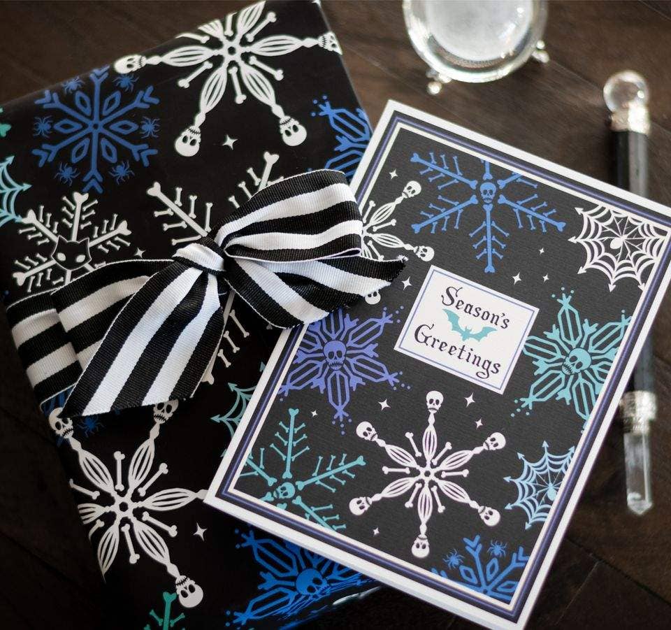 A gift-wrapped box and a matching greeting card laid on a dark wooden surface. The wrapping paper and card feature a winter snowflake and spiderweb pattern in black, white, blue, and light blue, with a black-and-white striped ribbon tied around the box.
