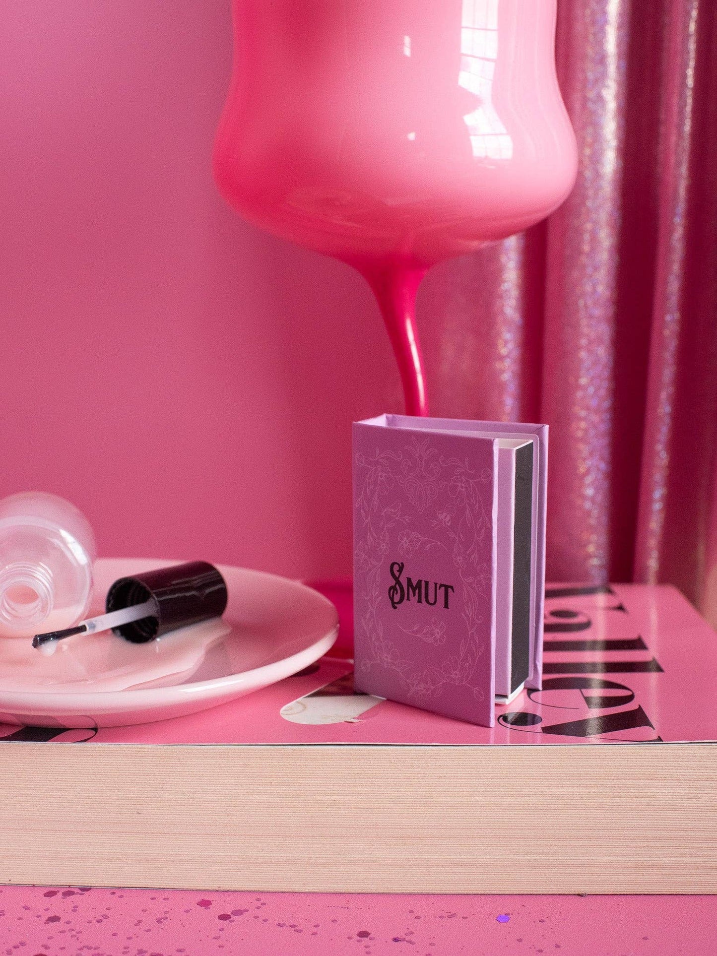 Pink bottle, nail polish, and book titled 'Smut' on a pink surface with a pink curtain.