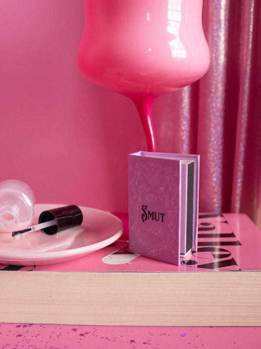 Pink bottle, nail polish, and book titled 'Smut' on a pink surface with a pink curtain.