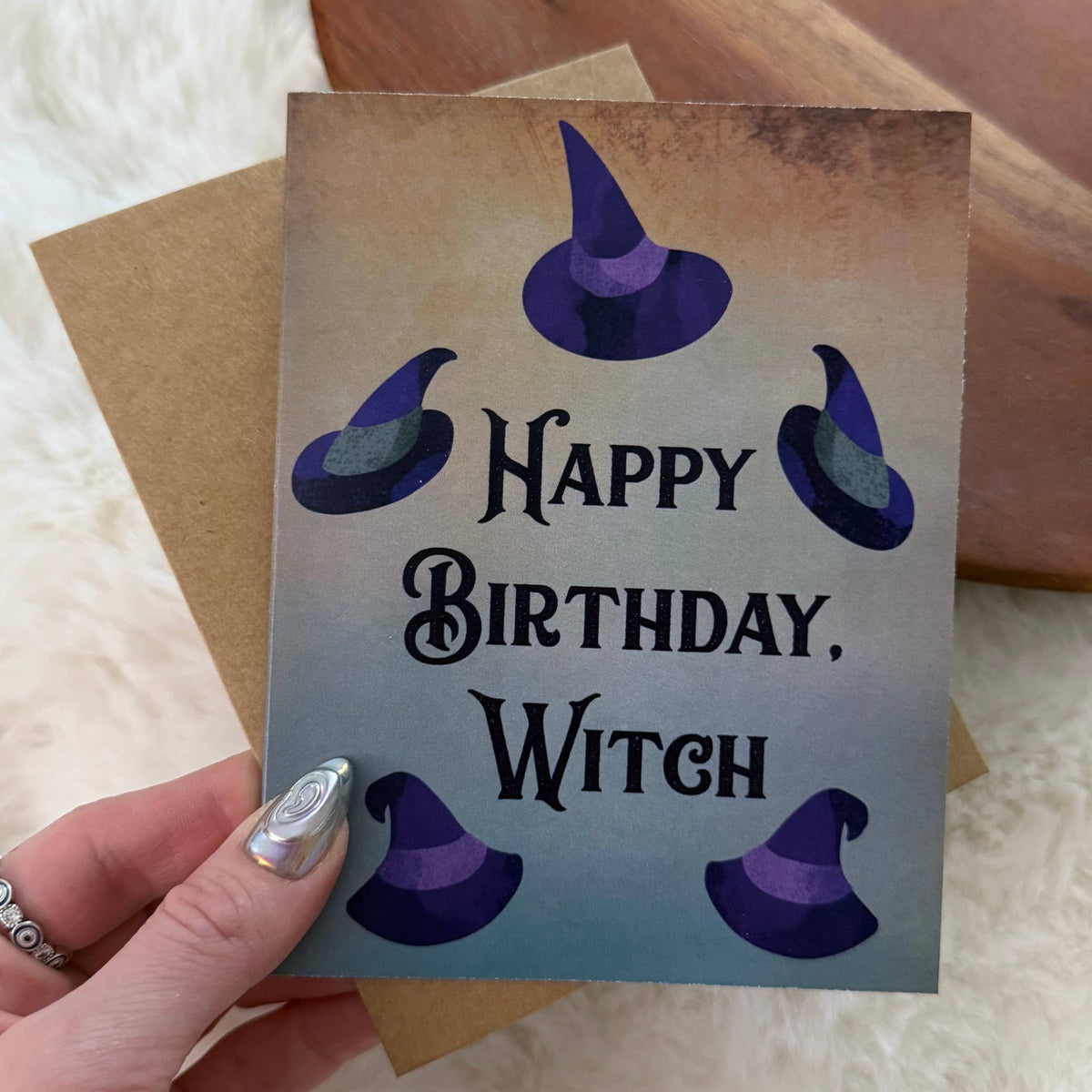 A hand holds a square birthday card that reads 'Happy Birthday, Witch' in black gothic lettering, decorated with five purple witch hats on a beige-to-blue gradient background. A brown envelope is partially visible behind it on a soft white surface.