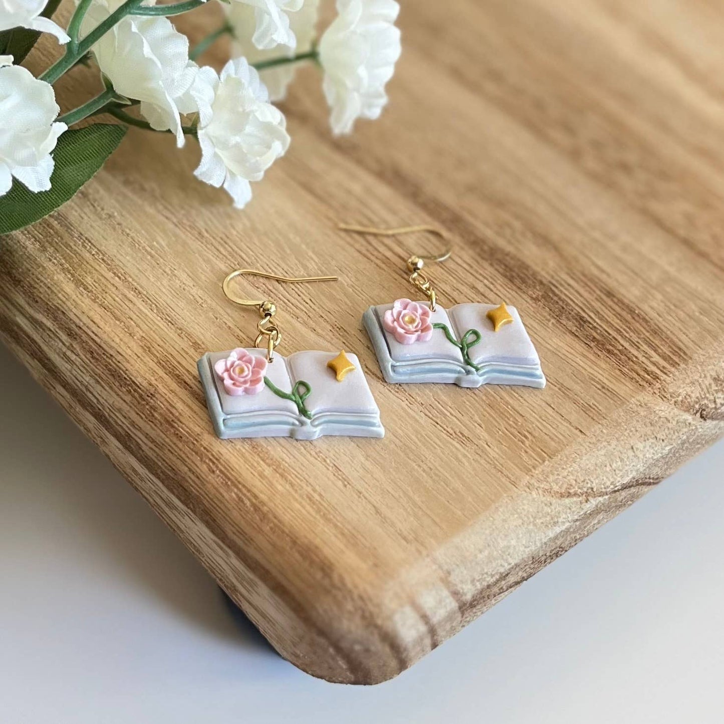 Book-shaped earrings with floral details on a wooden surface with flowers.