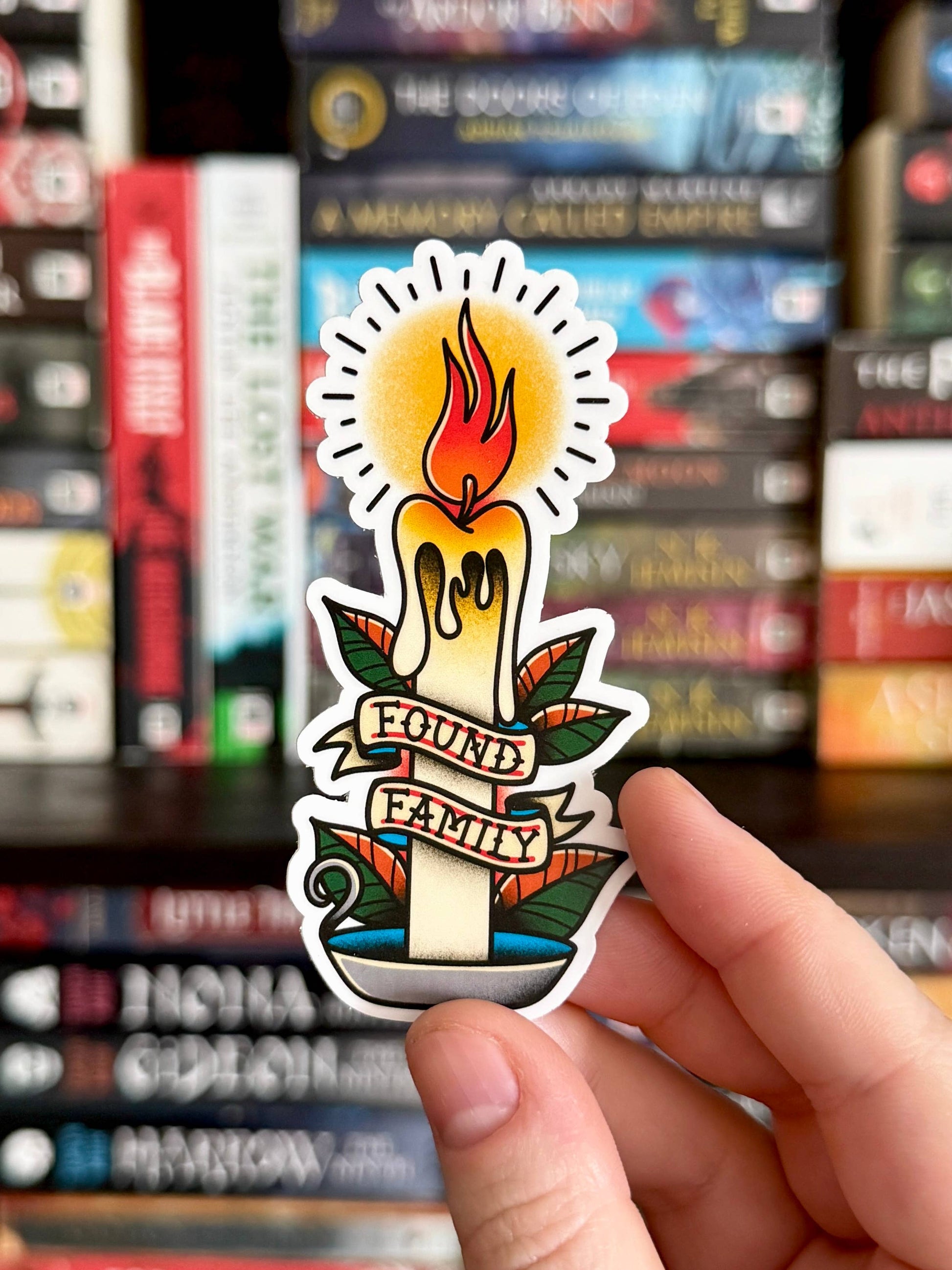 Sticker with a candle design and 'Found Family' text held in front of bookshelves.