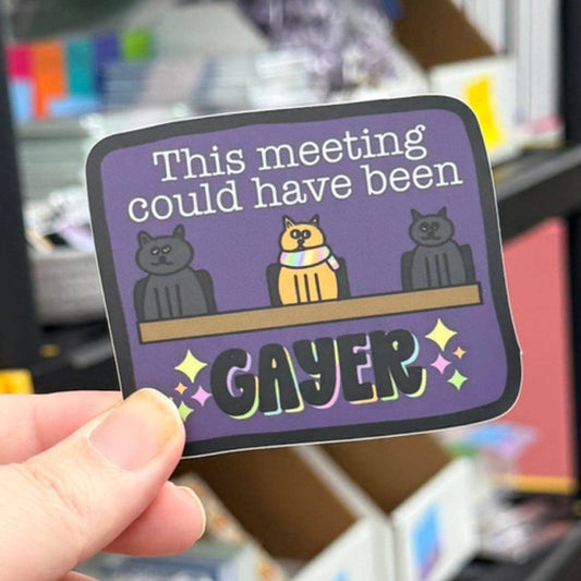 A hand holds a die-cut sticker with a purple background and cartoon cats sitting at a table. The sticker reads 'This meeting could have been GAYER' with colorful sparkles and rainbow accents.