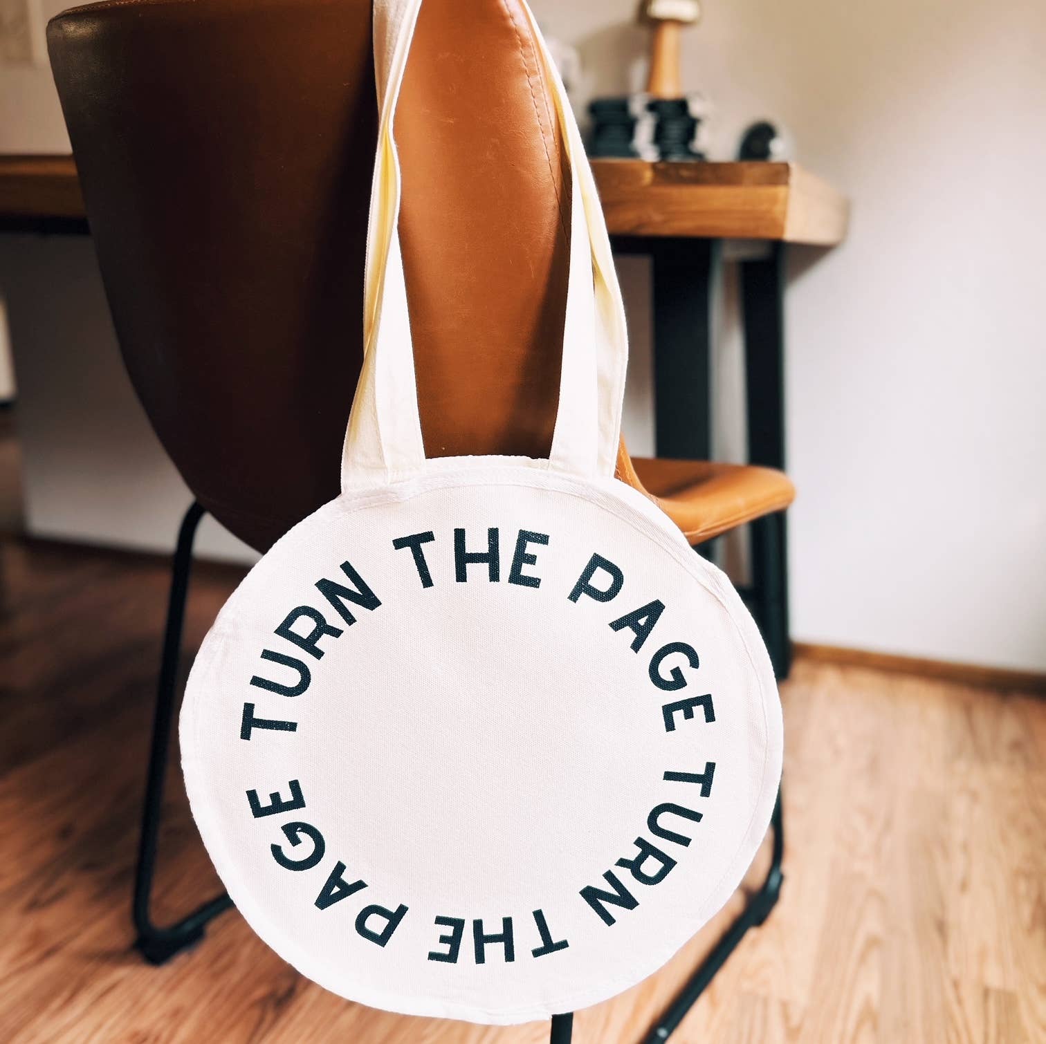 White tote bag with 'TURN THE PAGE' text hanging on a chair indoors.