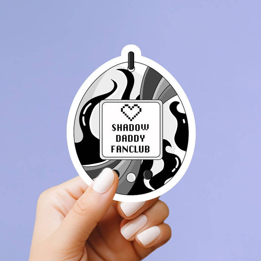 A hand with white manicured nails holds a round sticker or decal featuring a black, white, and gray swirled design with a central white box that reads 'SHADOW DADDY FANCLUB' and a small pixel heart. The background is a solid light lavender-blue color.