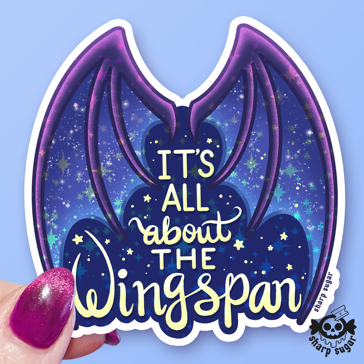 A die-cut vinyl sticker featuring purple bat wings on a starry blue night-sky background with the text 'IT'S ALL about THE Wingspan' in yellow and white. A hand with glittery pink nails holds the bottom-left corner of the sticker against a solid light blue background.