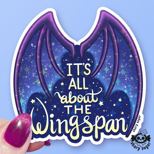 A die-cut vinyl sticker featuring purple bat wings on a starry blue night-sky background with the text 'IT'S ALL about THE Wingspan' in yellow and white. A hand with glittery pink nails holds the bottom-left corner of the sticker against a solid light blue background.