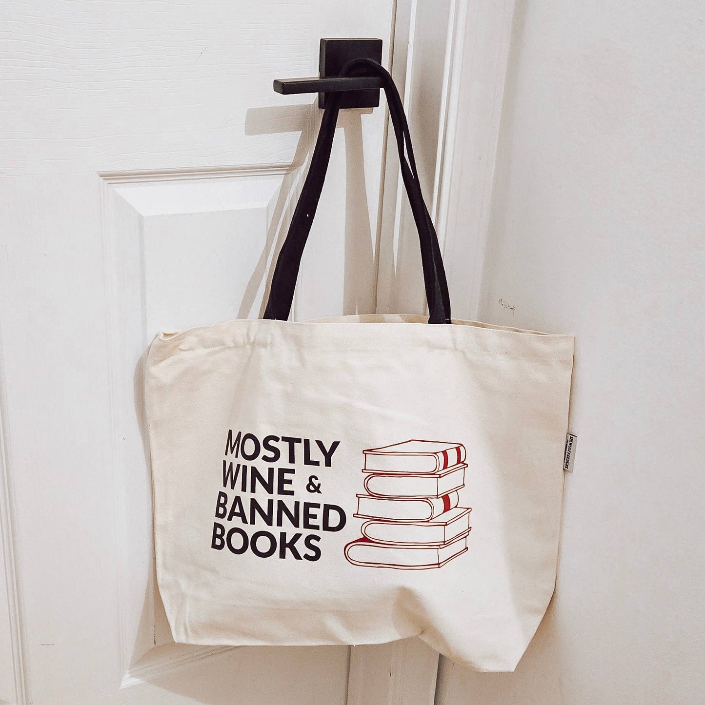 Tote bag with 'Mostly Wine & Banned Books' text and book graphic on a hook.