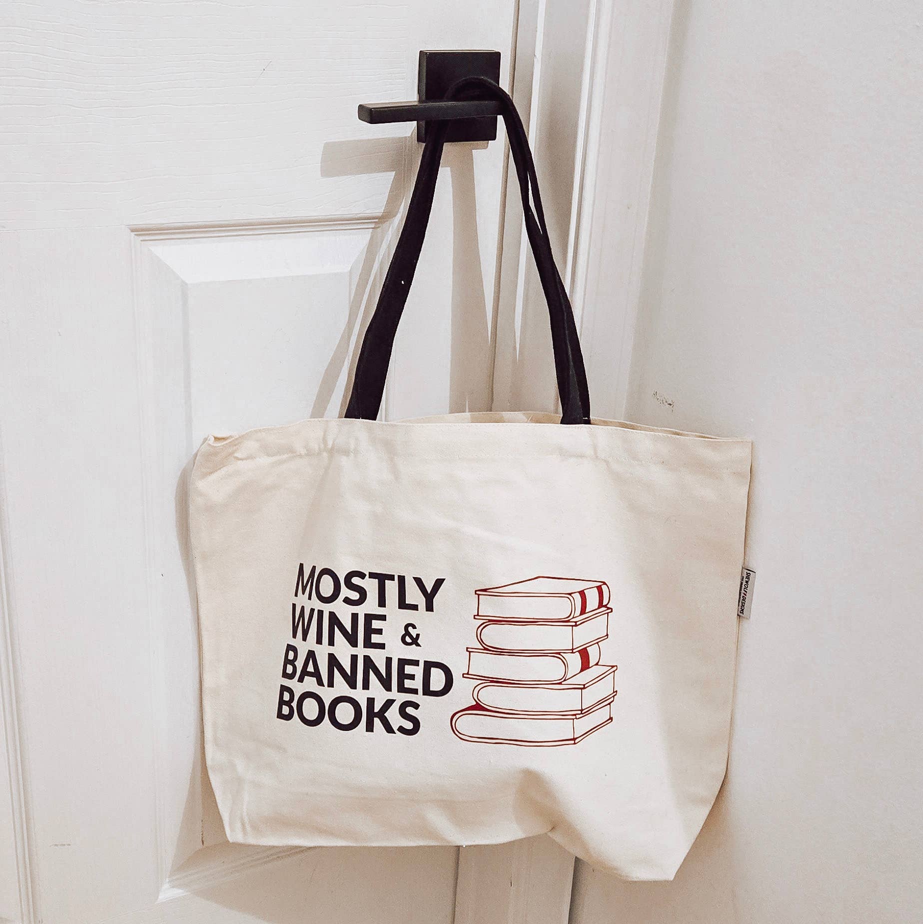 Tote bag with 'Mostly Wine & Banned Books' text and book graphic on a hook.