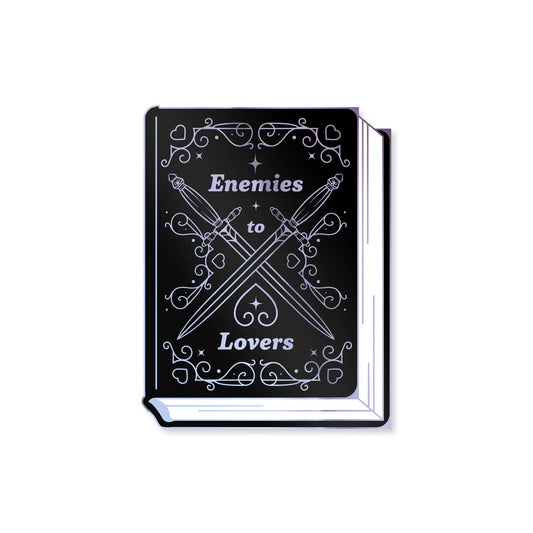 A black hardcover book or notebook with white gothic-style text reading 'Enemies to Lovers' and crossed swords with decorative flourishes on the cover, shown on a plain white background.