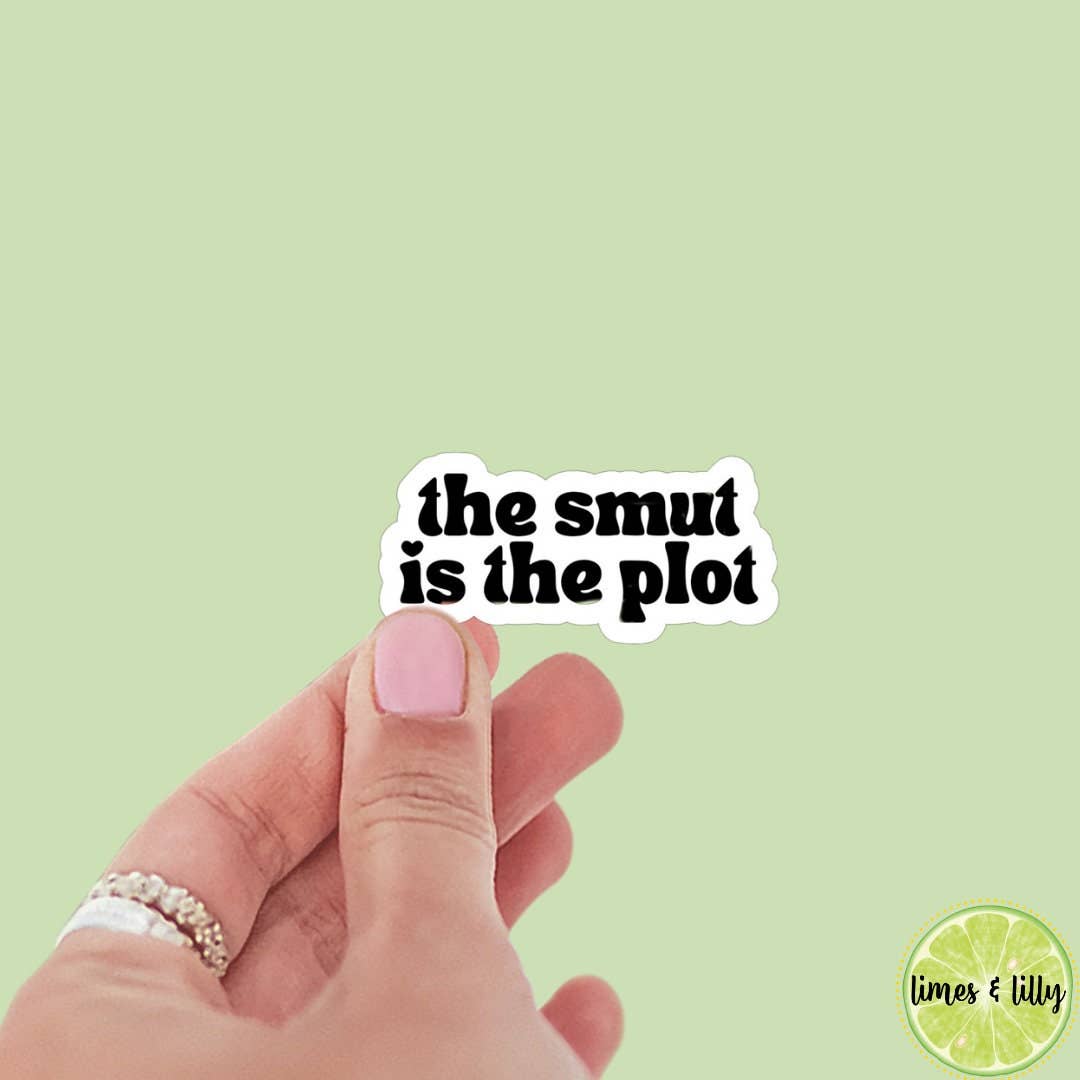 Hand holding a sticker with 'the smut is the plot' text on a light green background