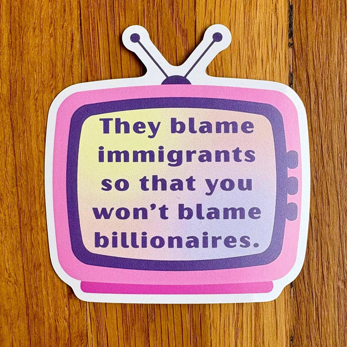 A die-cut sticker shaped like a retro television with the text 'They blame immigrants so that you won't blame billionaires.' in purple lettering on a pink and lavender gradient face, placed on a wooden surface.