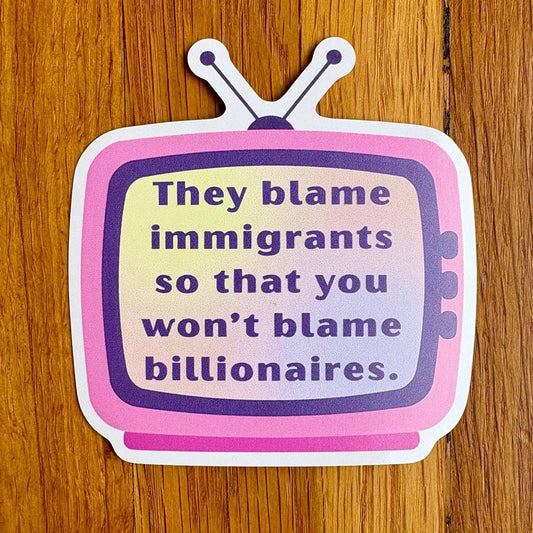 A die-cut sticker shaped like a retro television with the text 'They blame immigrants so that you won't blame billionaires.' in purple lettering on a pink and lavender gradient face, placed on a wooden surface.