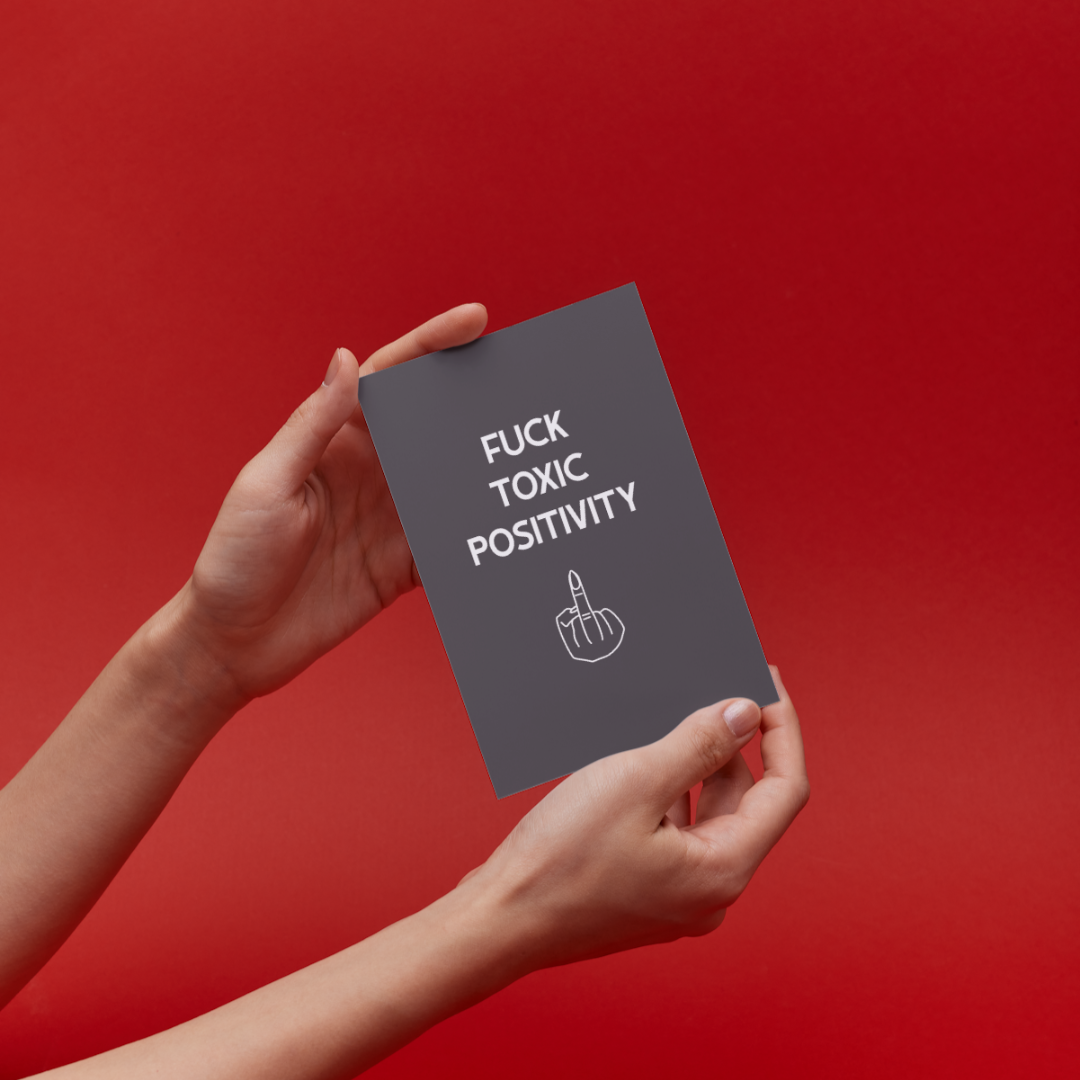 Hands holding a book titled 'F*ck Toxic Positivity' against a red background