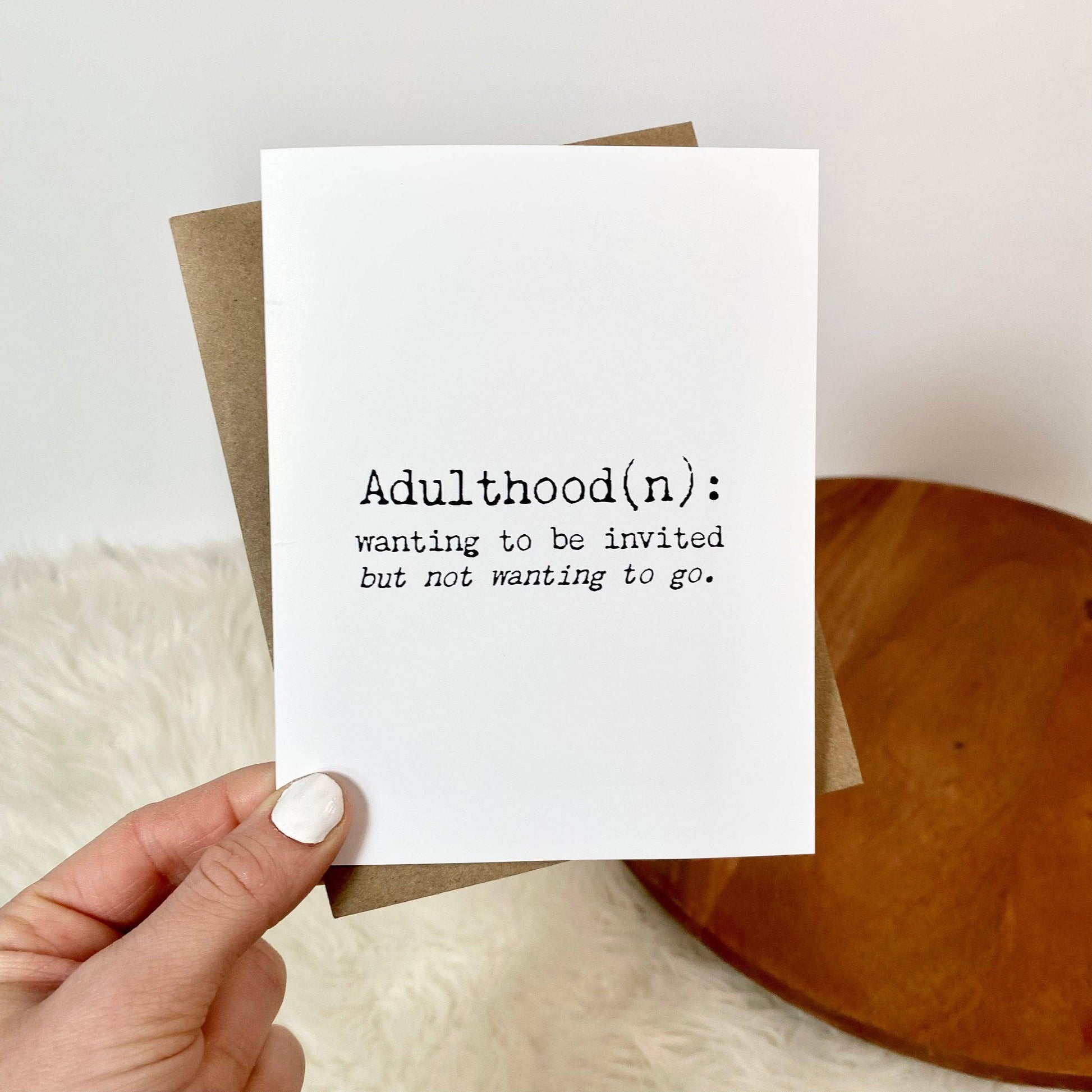 A hand holds a white greeting card with black printed text reading 'Adulthood(n): wanting to be invited but not wanting to go.' A brown envelope is partially visible behind the card, set against a soft white faux-fur surface with a round wooden tray in the background.