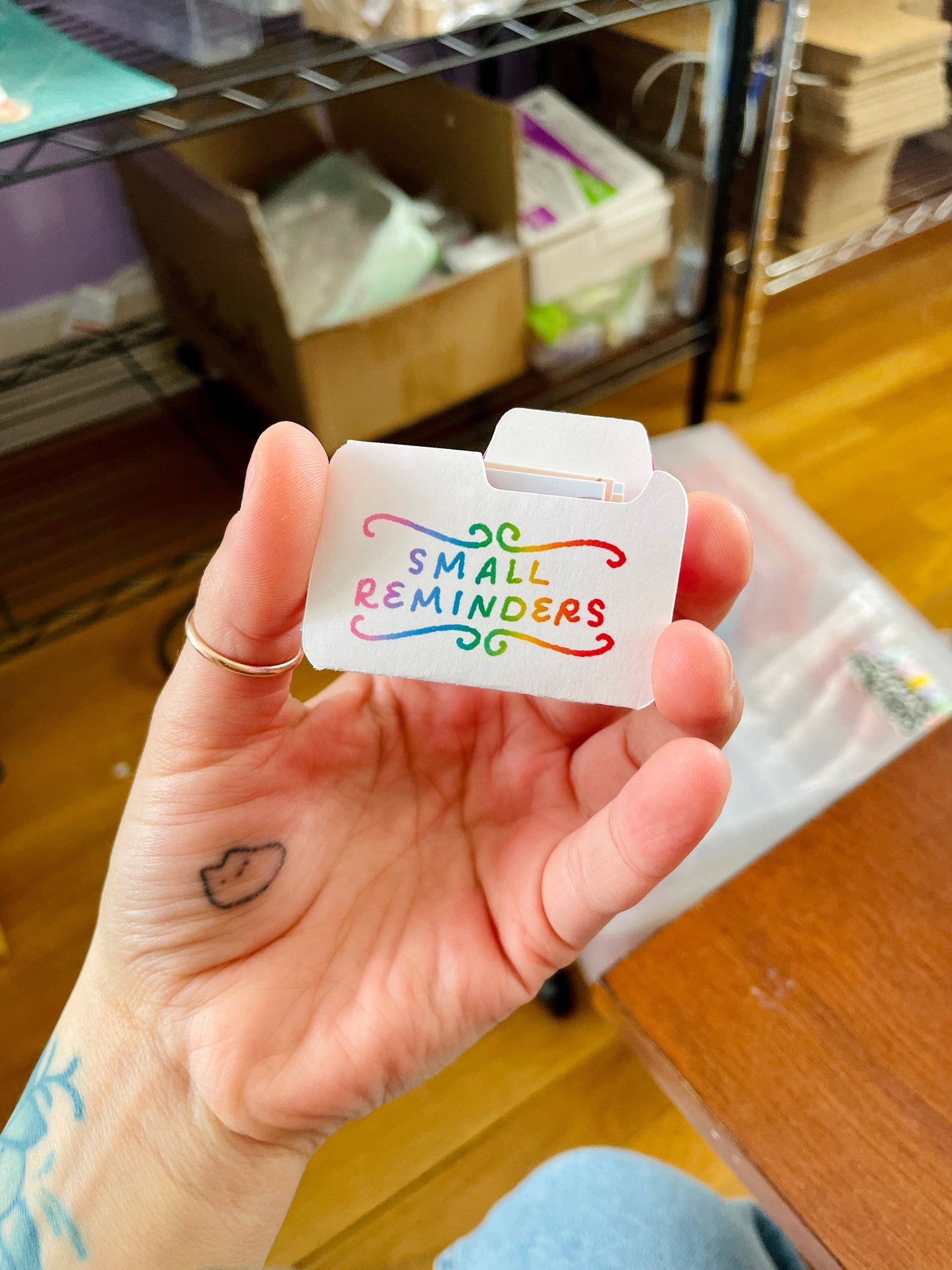 Hand holding a small white card with colorful text 'Small Reminders' in an indoor setting.