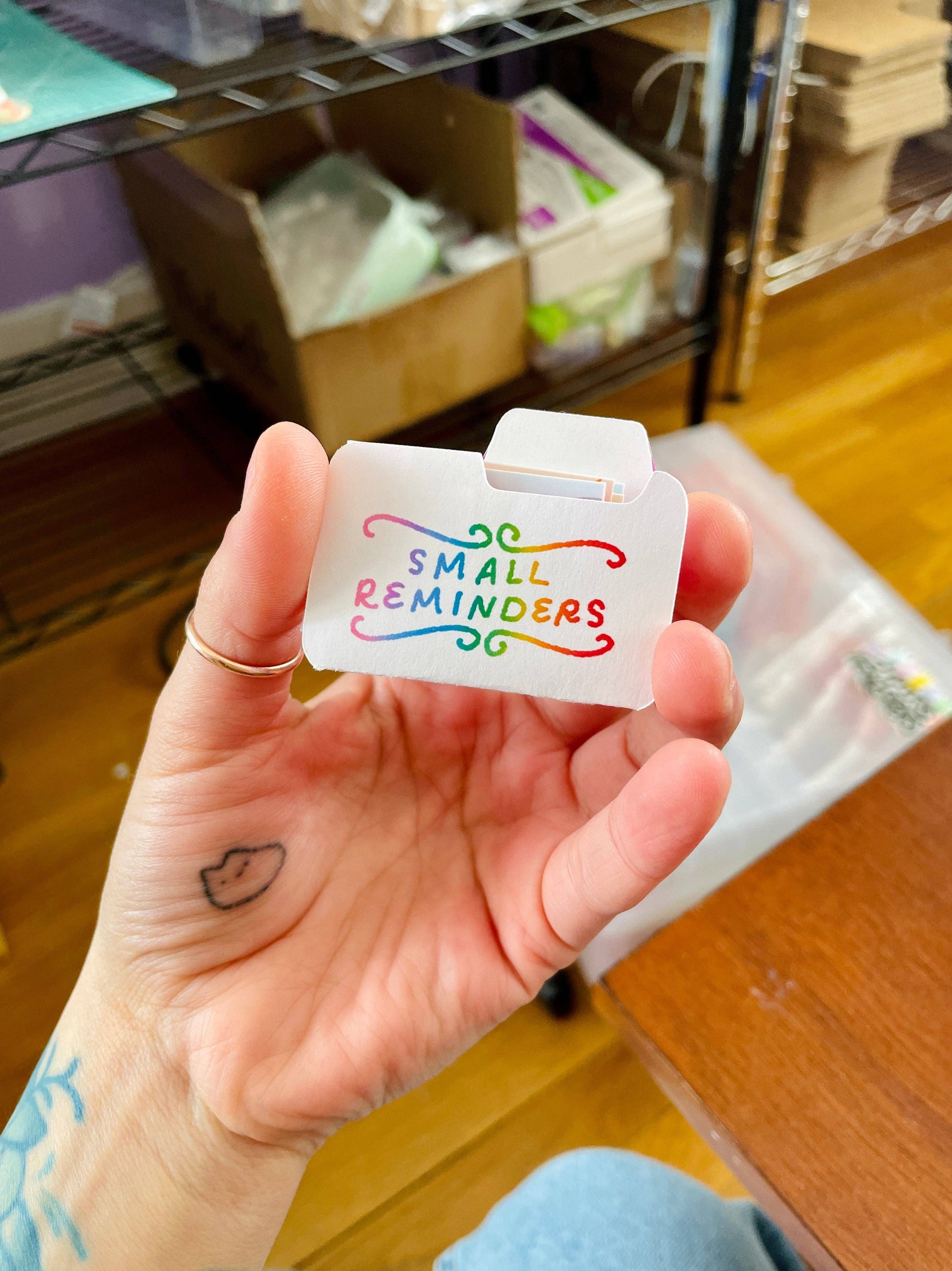 Hand holding a small white card with colorful text 'Small Reminders' in an indoor setting.