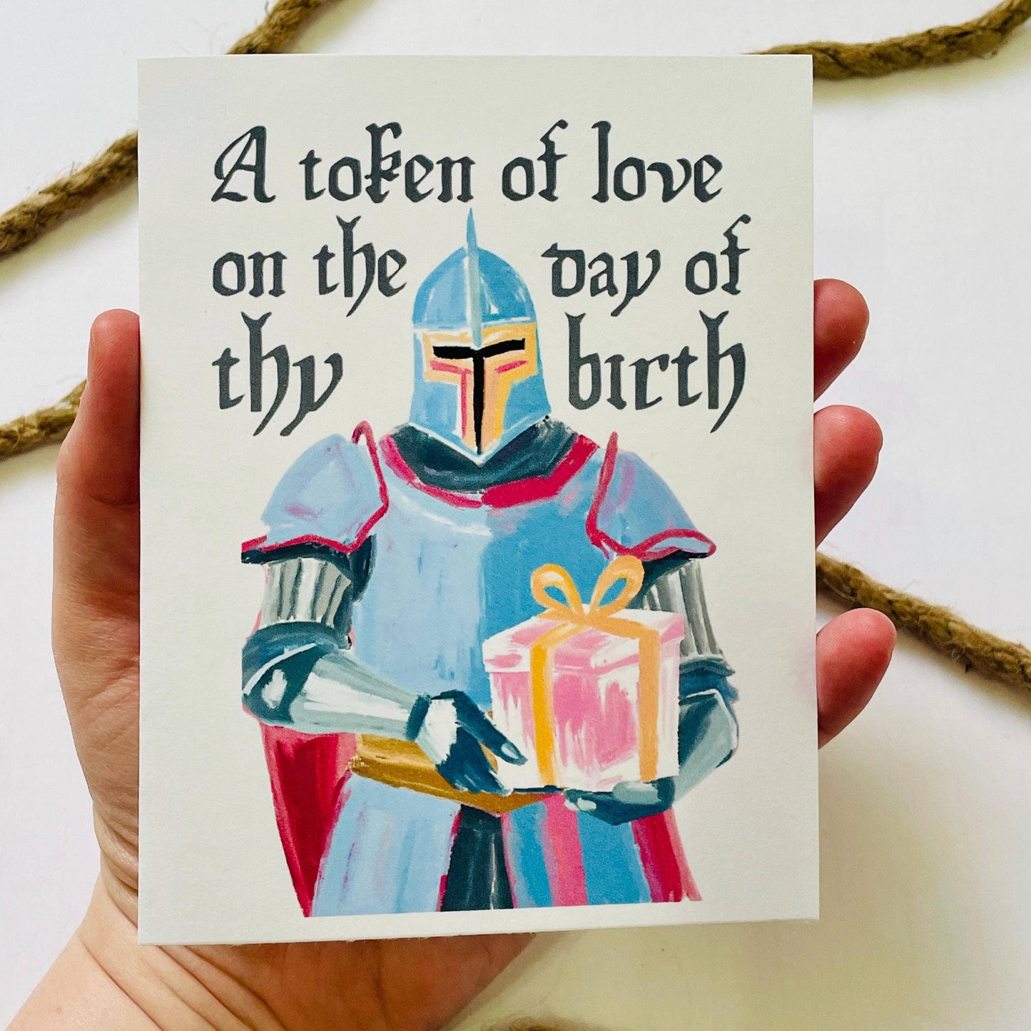 Card with a knight holding a gift and text 'A token of love on the day of thy birth' held by a hand.