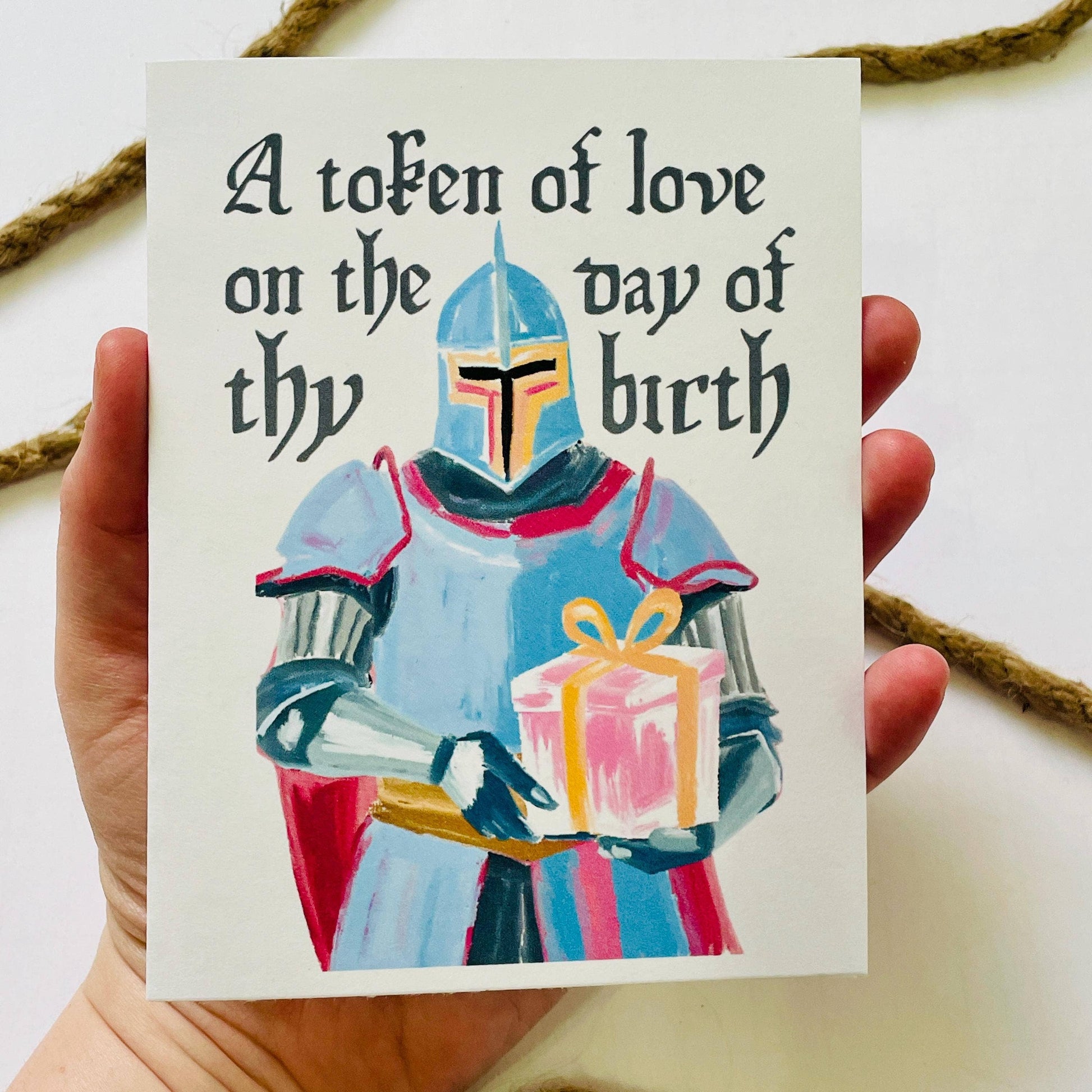Card with a knight holding a gift and text 'A token of love on the day of thy birth' held by a hand.