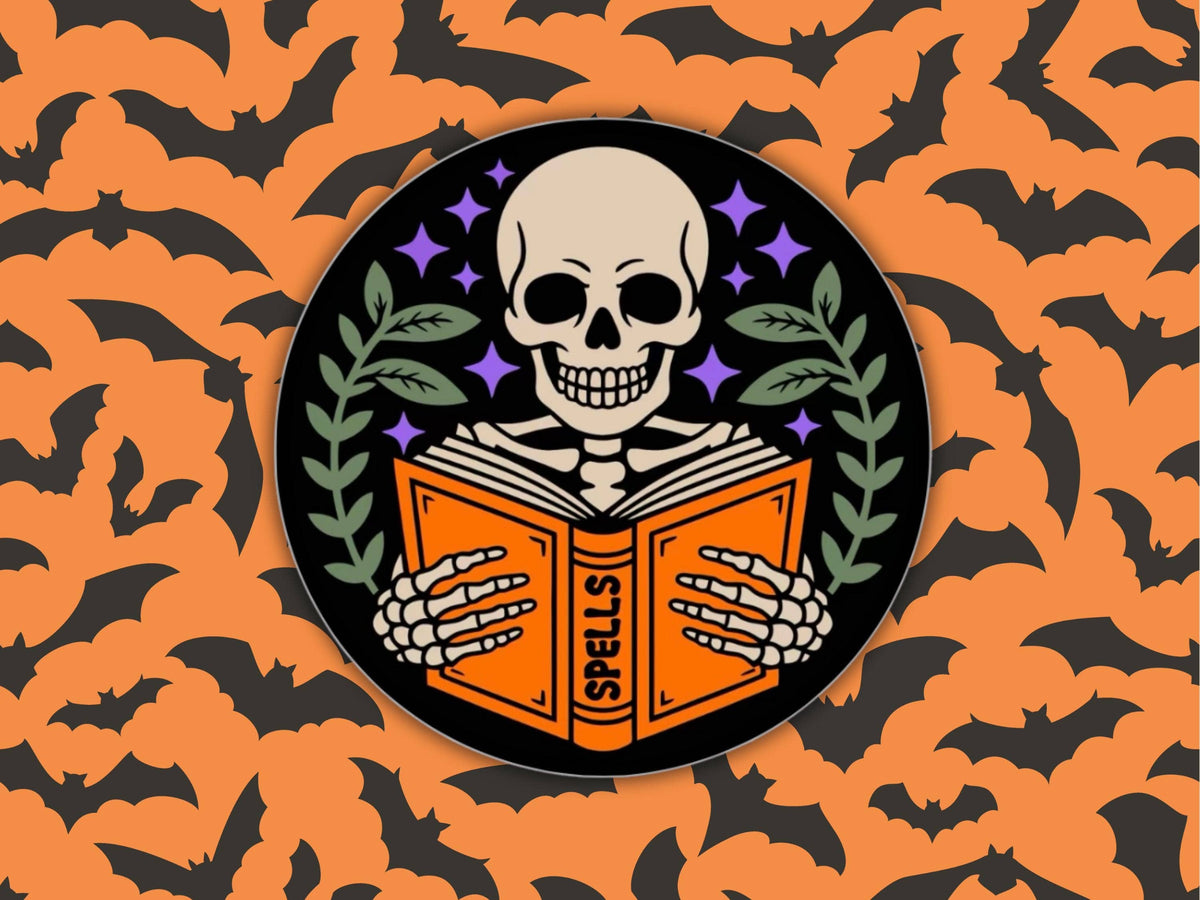 A circular Halloween-themed graphic featuring a skull skeleton holding an orange book labeled 'SPELLS', set against a black circle with purple stars and green leaves. The design is centered on an orange background patterned with black bats.