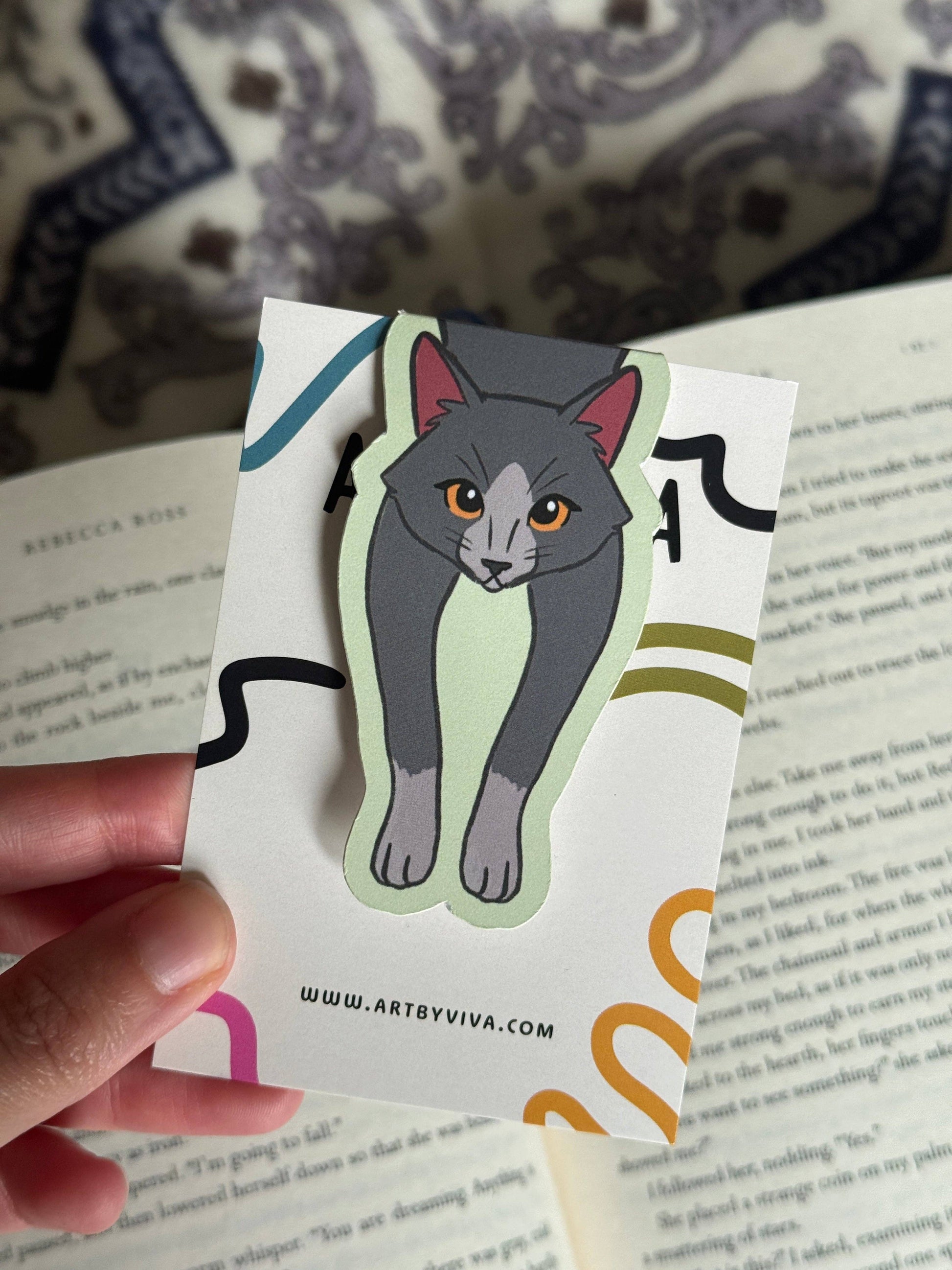 Sticker of a cat held over a book with 'www.artbyviva.com' printed on it.