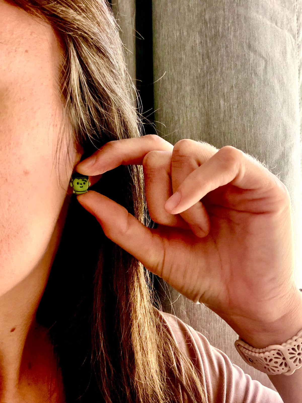 A close-up lifestyle shot of a woman wearing a small green beaded earring, holding her hand near her ear to show the earring's scale, with a neutral gray curtain in the background.