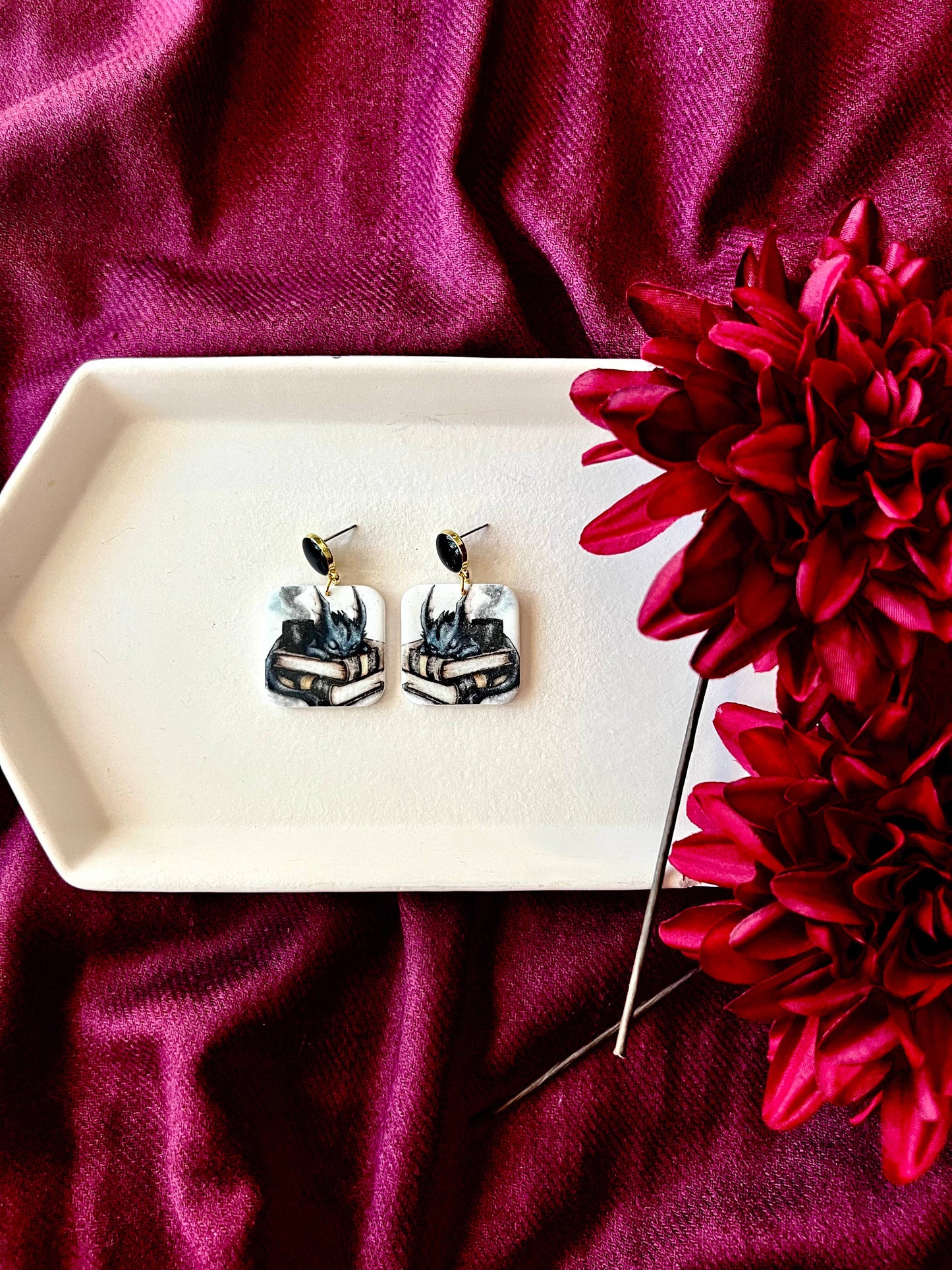 Pair of earrings on a white dish with a red flower on a purple fabric background