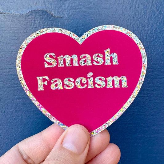 A hand holds a heart-shaped sticker with a bright pink background and a glittery multicolor border. The sticker features the text 'Smash Fascism' in white glitter letters against a dark blue textured background.