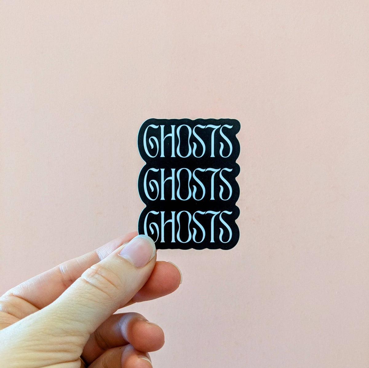 A hand holds a small rectangular sticker with the word 'GHOSTS' repeated three times in a stylized light blue font on a black background, against a soft pink wall.