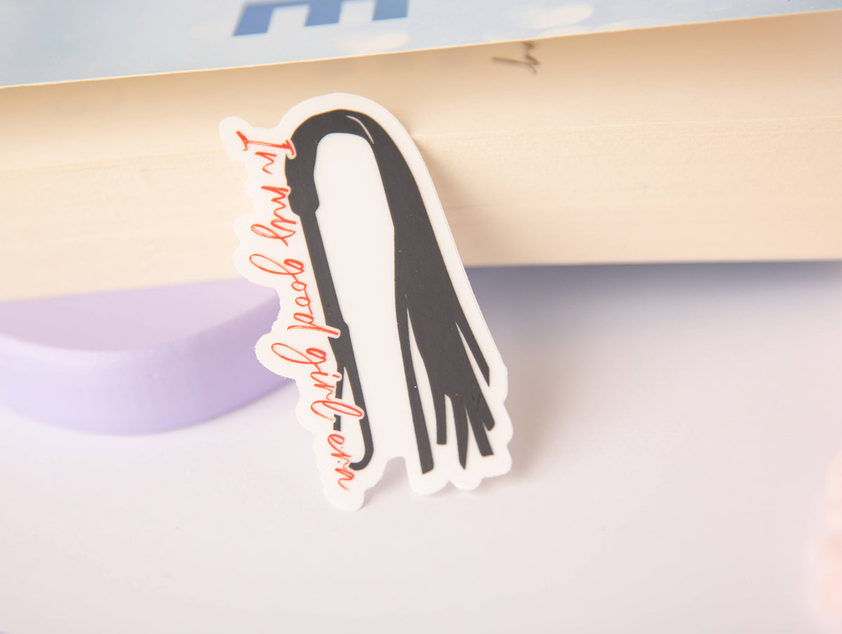A die-cut sticker featuring a black hair silhouette with red cursive text reading 'I'm my good girl team' on a white background, photographed leaning against a book.