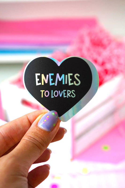 A hand with lavender glitter nail polish holds a heart-shaped black sticker that reads 'ENEMIES TO LOVERS' in pastel lettering. The background is a soft-focus pink and white box.