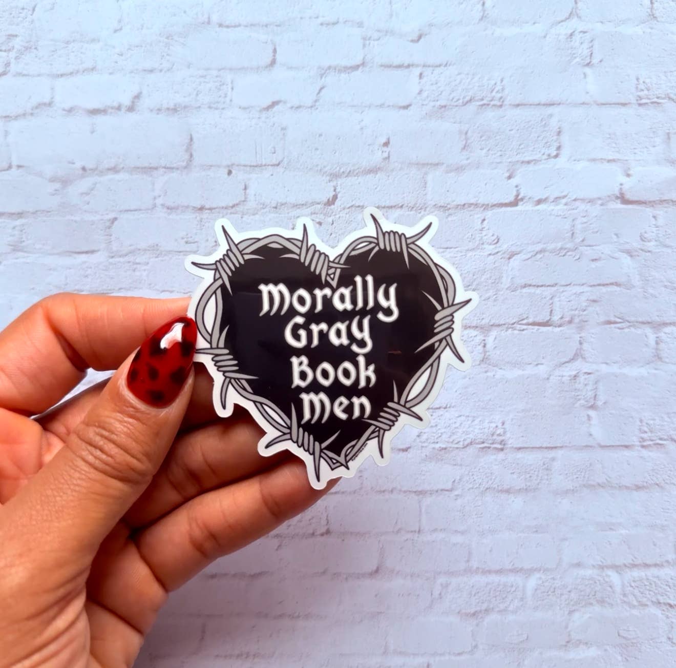 Hand holding a 'Morally Gray Book Men' sticker against a light blue brick wall.