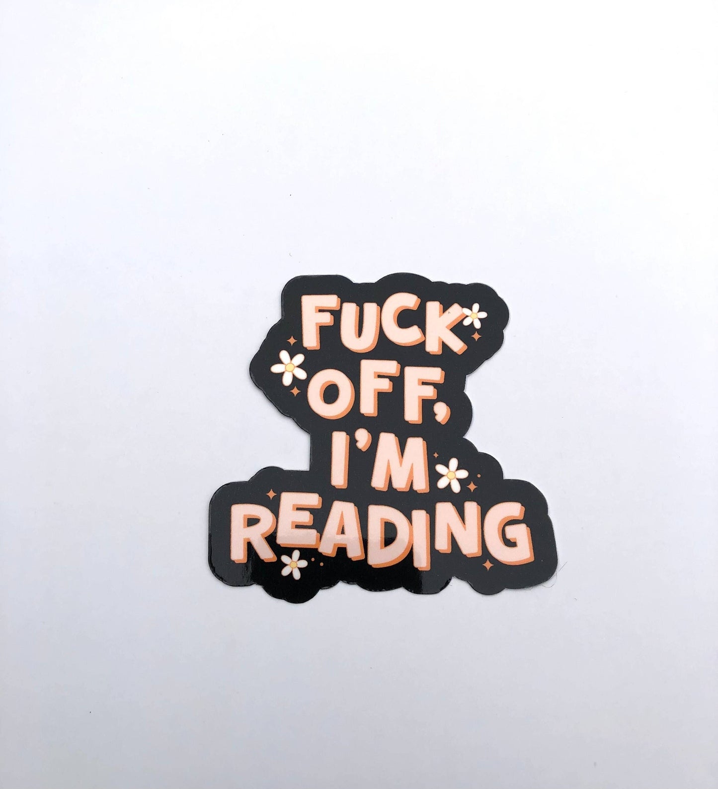 A die-cut sticker with the text 'FUCK OFF, I'M READING' in peach-colored bubble letters on a black background, accented with small white starburst shapes, shown on a plain white surface.