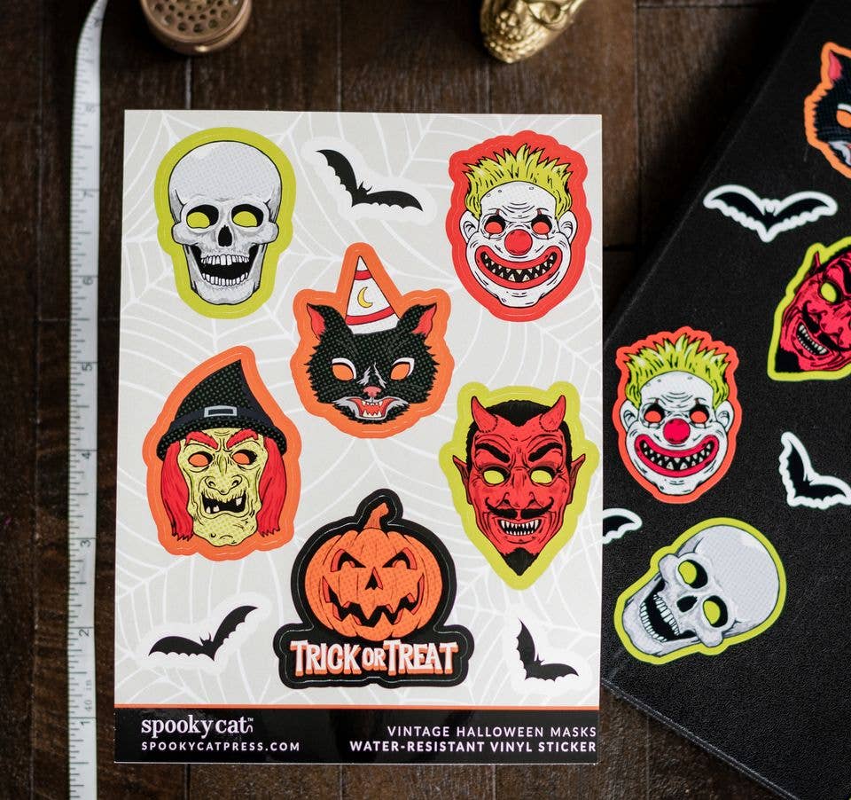 A sheet of vintage-style Halloween vinyl stickers displayed on a dark wooden surface next to a ruler and a black sticker roll. The stickers feature classic spooky faces and motifs including a skull, clown, witch, devil, cat in a hat, jack-o'-lantern, bats, and spiderwebs.
