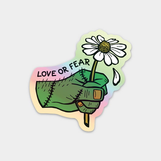 A die-cut sticker featuring a stitched green hand holding a white daisy with the text 'LOVE OR FEAR' on a pastel iridescent background, shown on a light gray studio backdrop.