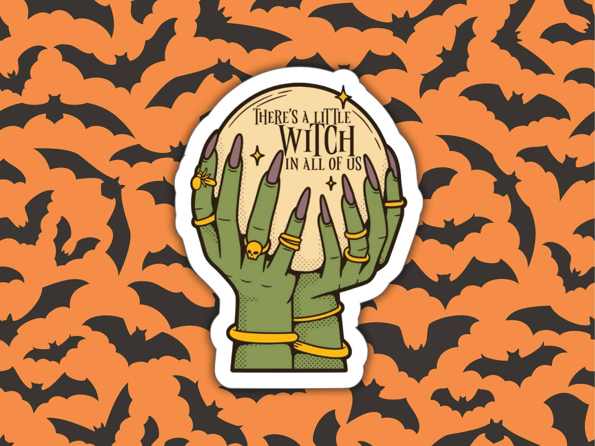 A die-cut sticker featuring two green skeletal hands holding a glowing moon with the text 'There's a little witch in all of us,' set against an orange and black bat-patterned background.
