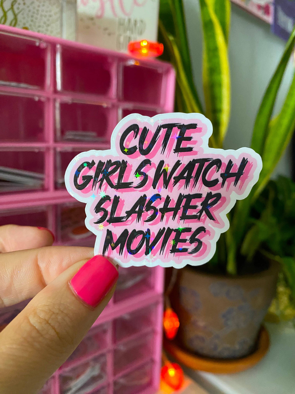 A hand with bright pink nail polish holds a die-cut sticker that reads 'CUTE GIRLS WATCH SLASHER MOVIES' in black text with a pink and white outline. The background shows a pink organizer box and a green potted plant.