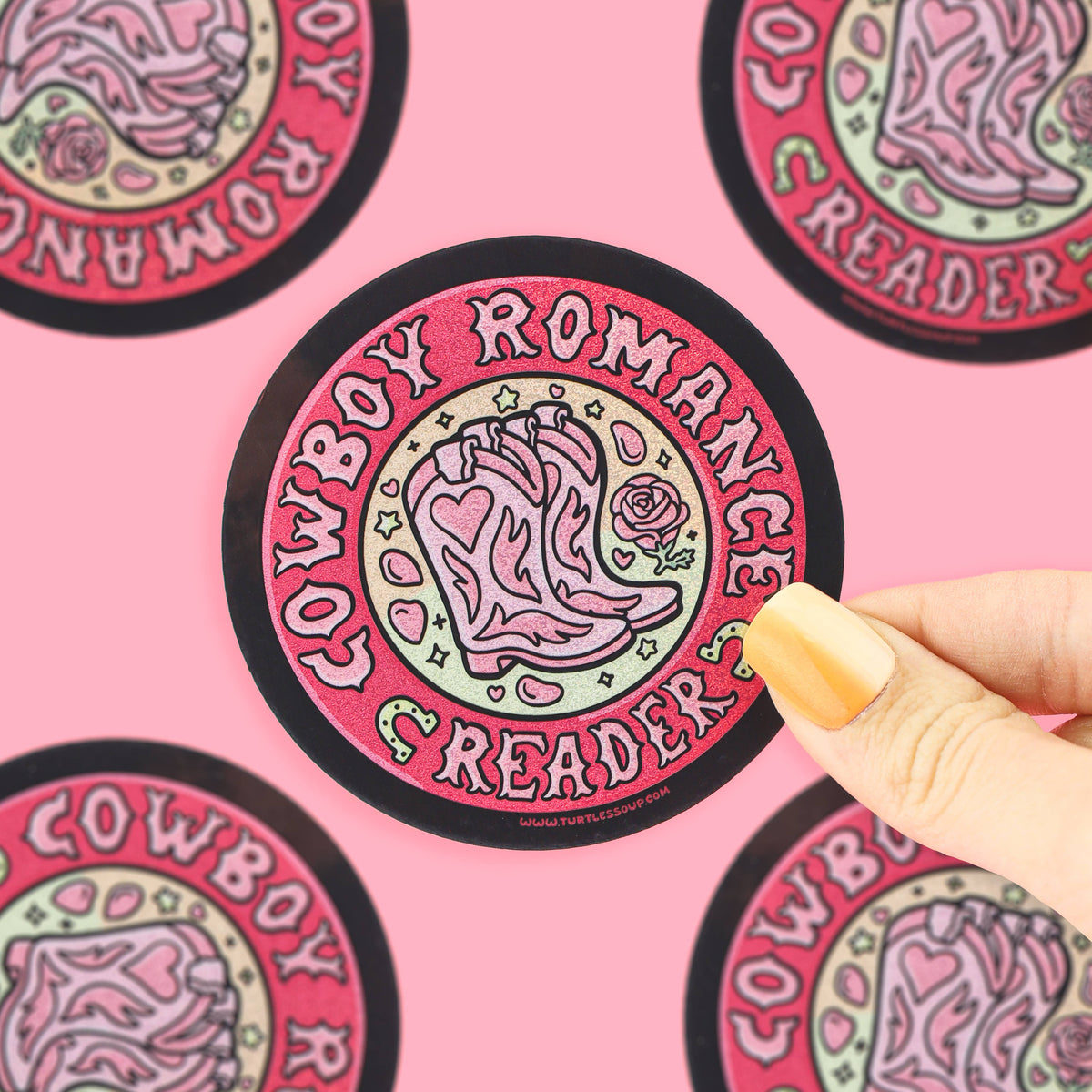 A hand with yellow nails holds a round sticker featuring a pink cowboy boot and the words 'COWBOY ROMANTICS READER' on a pink background with other matching stickers partially visible.