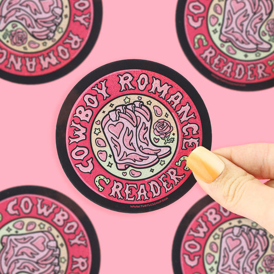 A hand with yellow nails holds a round sticker featuring a pink cowboy boot and the words 'COWBOY ROMANTICS READER' on a pink background with other matching stickers partially visible.
