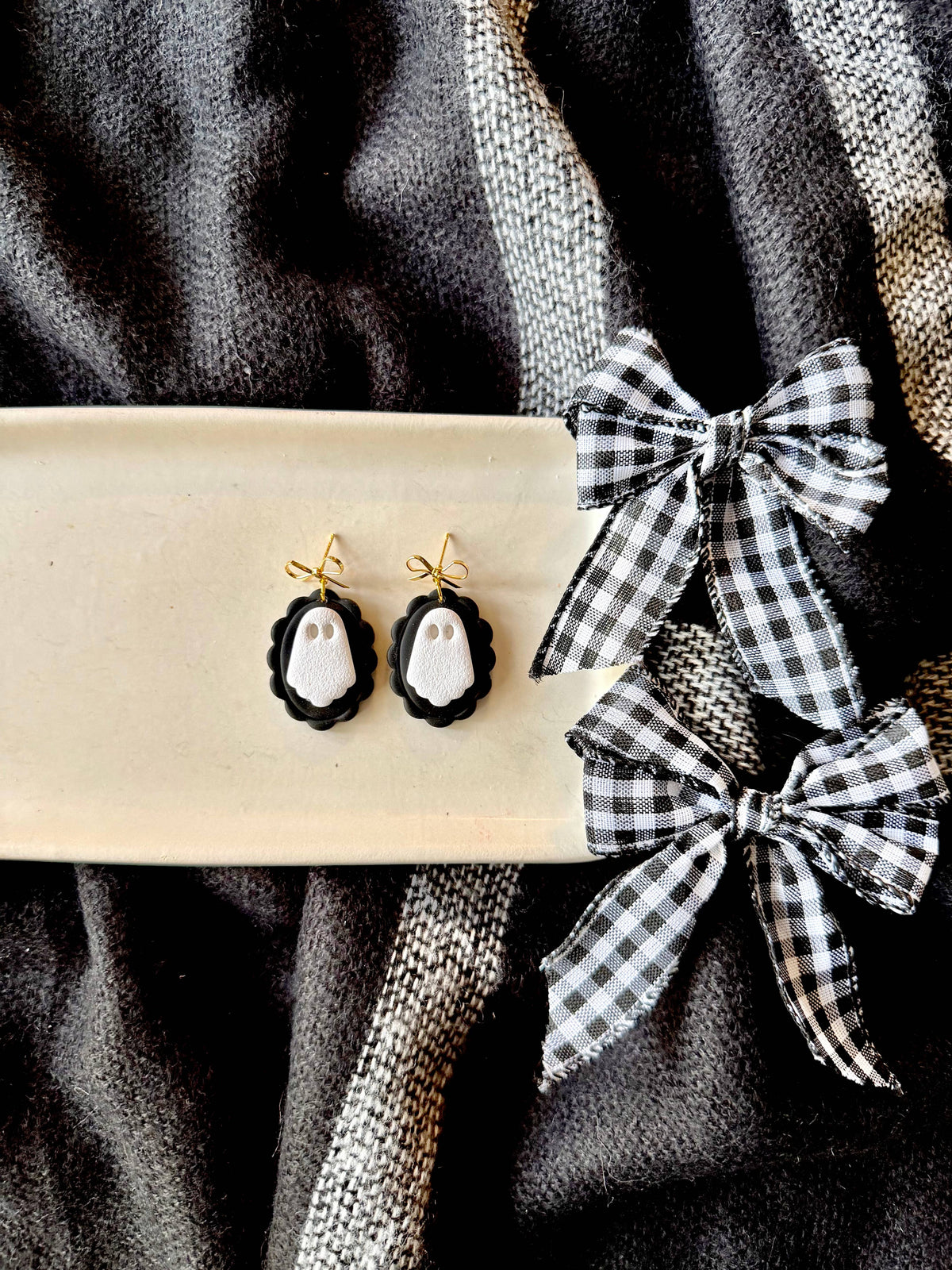 Black and white ghost-themed drop earrings displayed on a cream-colored rectangular tray, next to two black and white gingham ribbon bows on a dark textured fabric background.